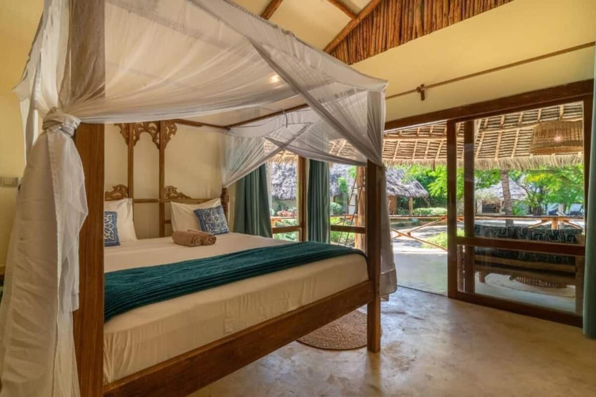 A four-poster bed with a canopy, a wooden headboard, and a view of a tropical garden through large glass doors.
