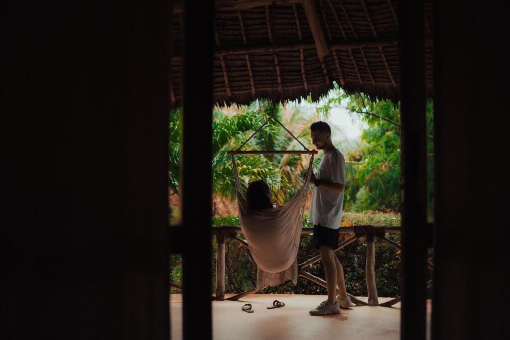 A hammock suspended between two wooden posts, a person standing nearby, and a thatched roof structure.