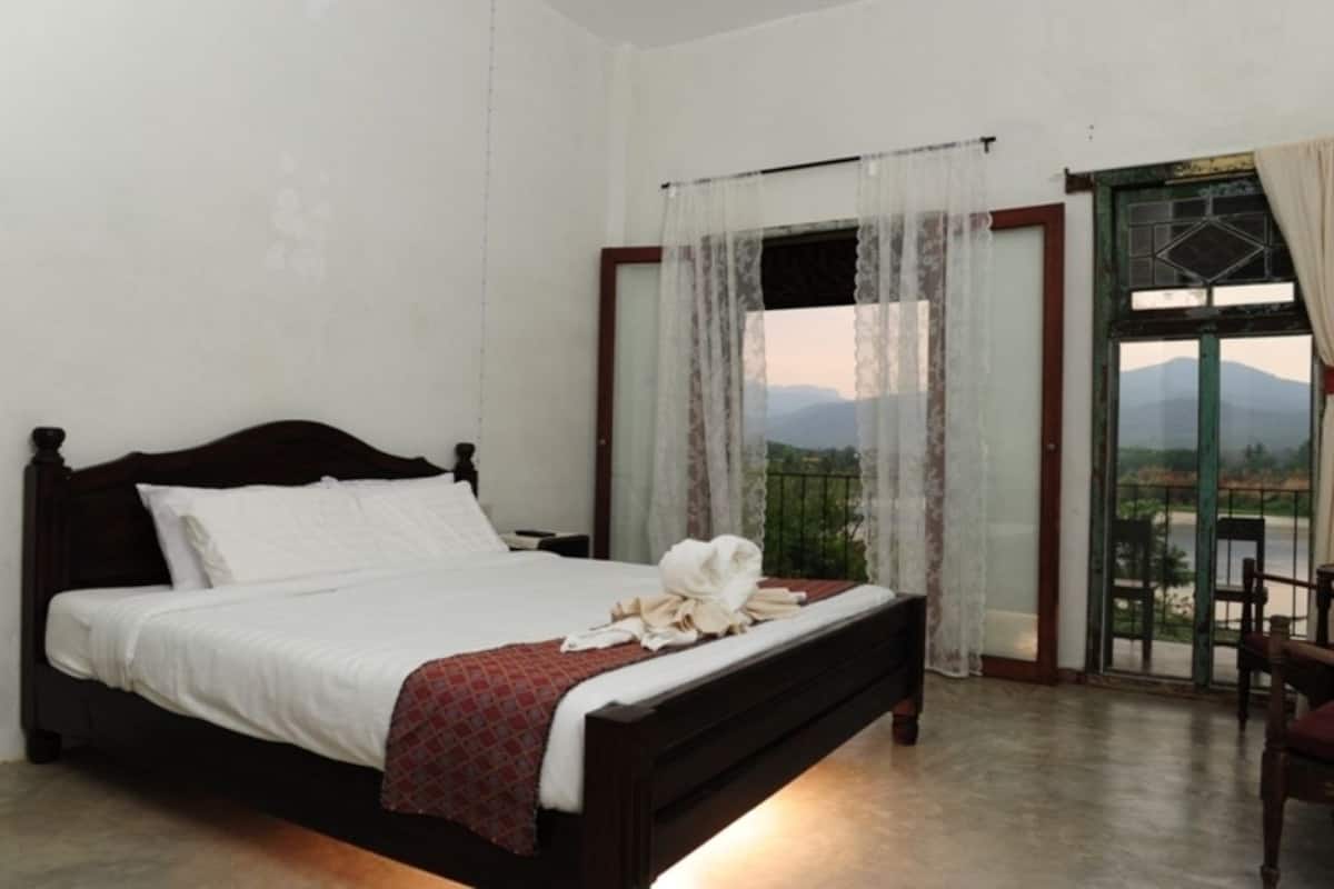 A single bed with a wooden frame, white bedding, and a red patterned throw. A white towel is placed on the bed. The room has a balcony with a view of mountains and a river.