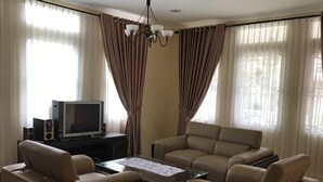 A living room with beige sofas, a coffee table, a TV, and curtains. - Villa Sofia Kota Bunga (Cipanas)