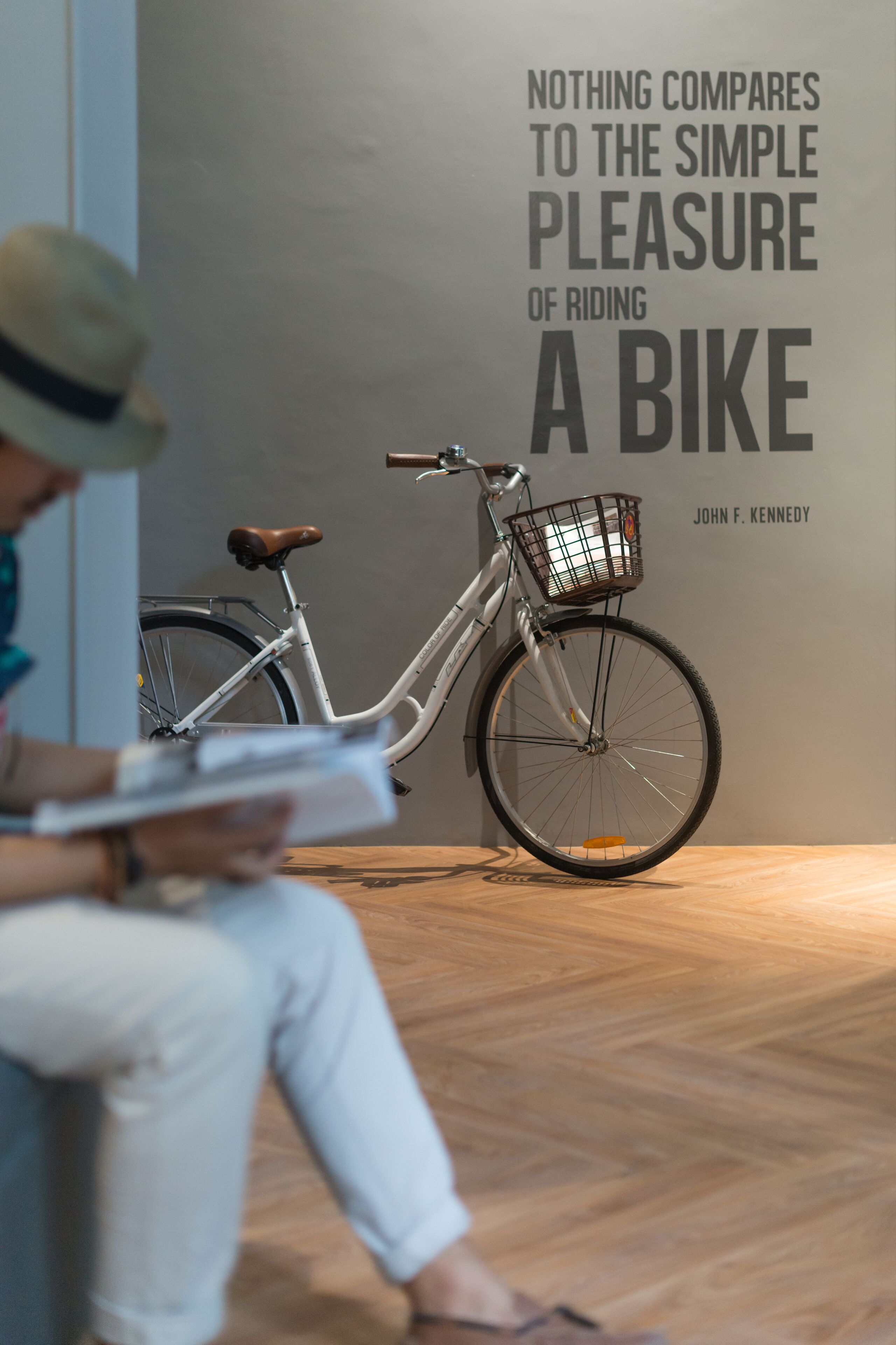 Una persona leyendo un libro junto a una bicicleta blanca con cesto, en una habitación con suelo de madera y una pared con una cita sobre andar en bicicleta.