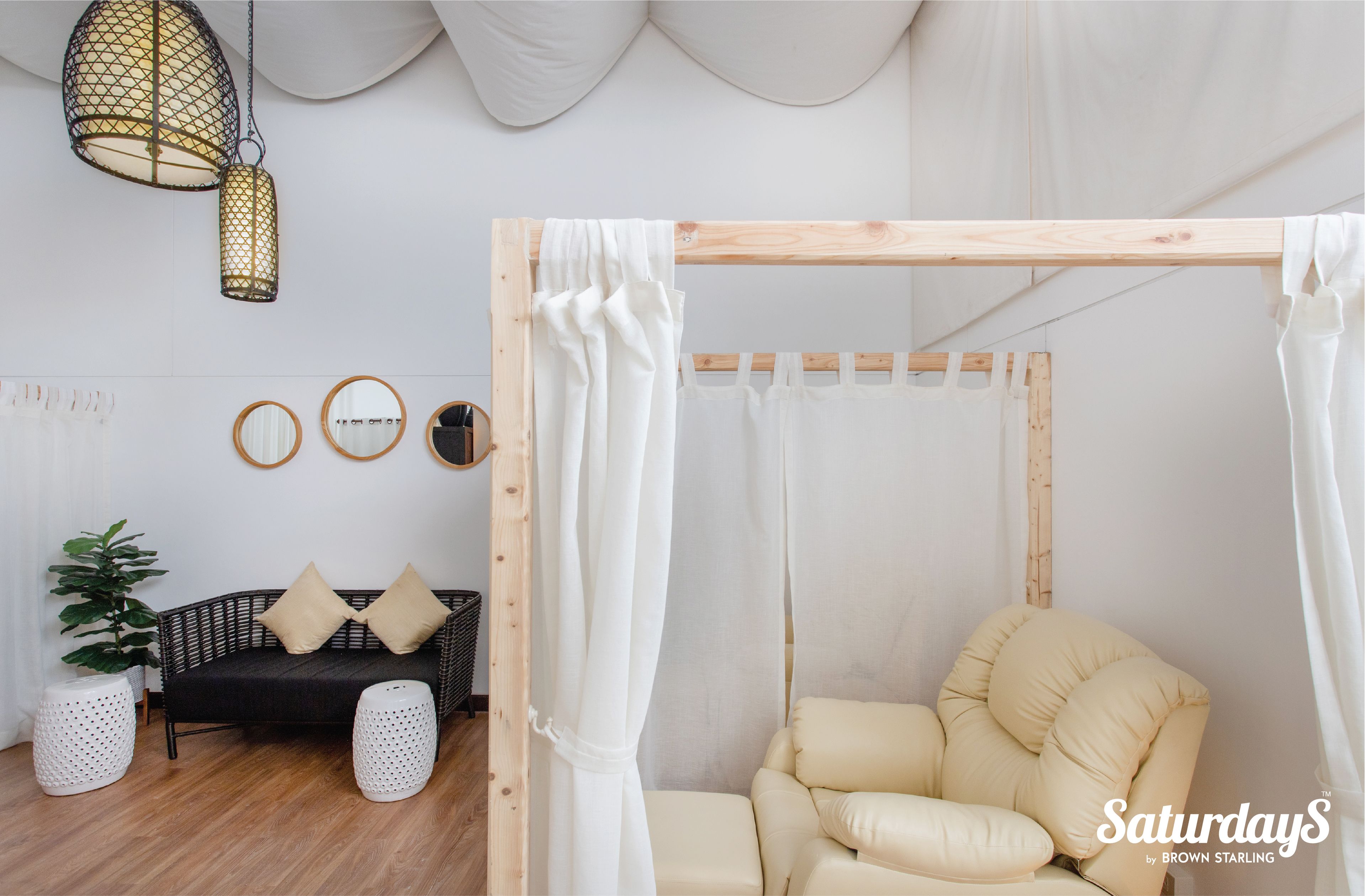 A room with a wooden bed frame, a white canopy, a beige armchair, a black sofa, and a potted plant.
