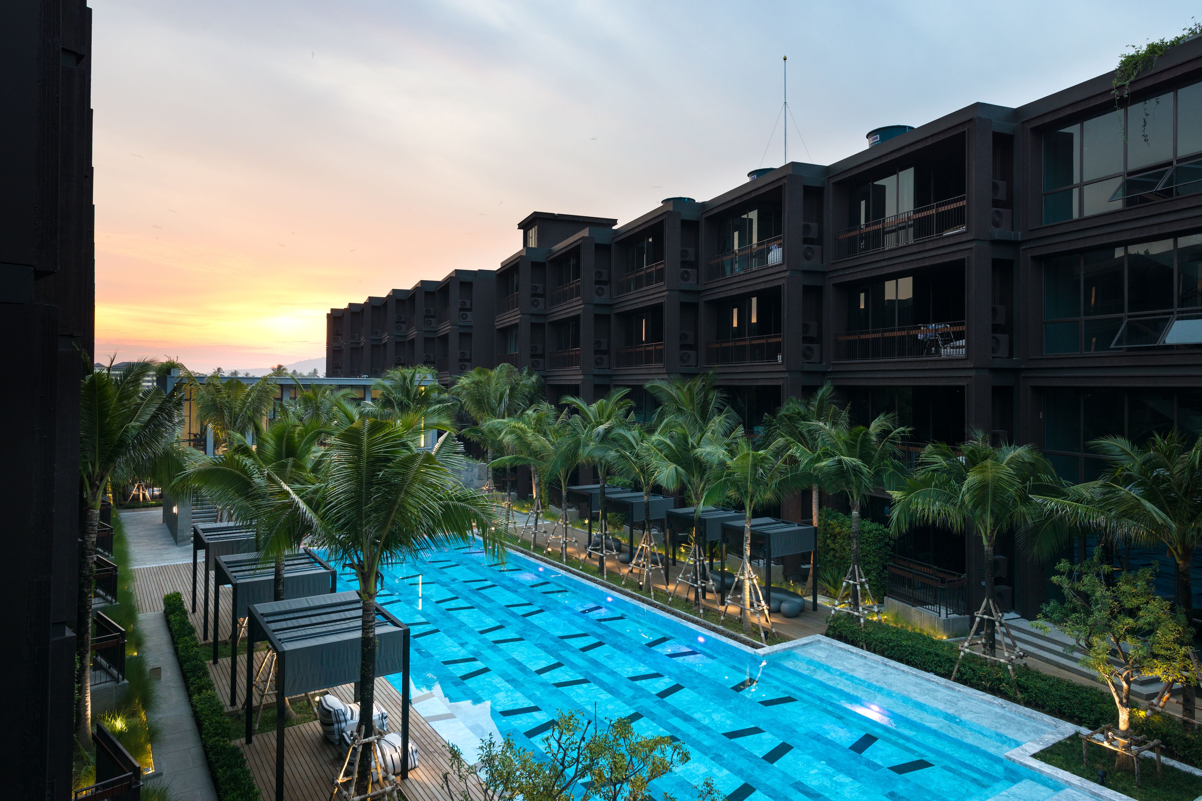 A modern hotel with a large outdoor swimming pool, surrounded by palm trees and a sunset view.