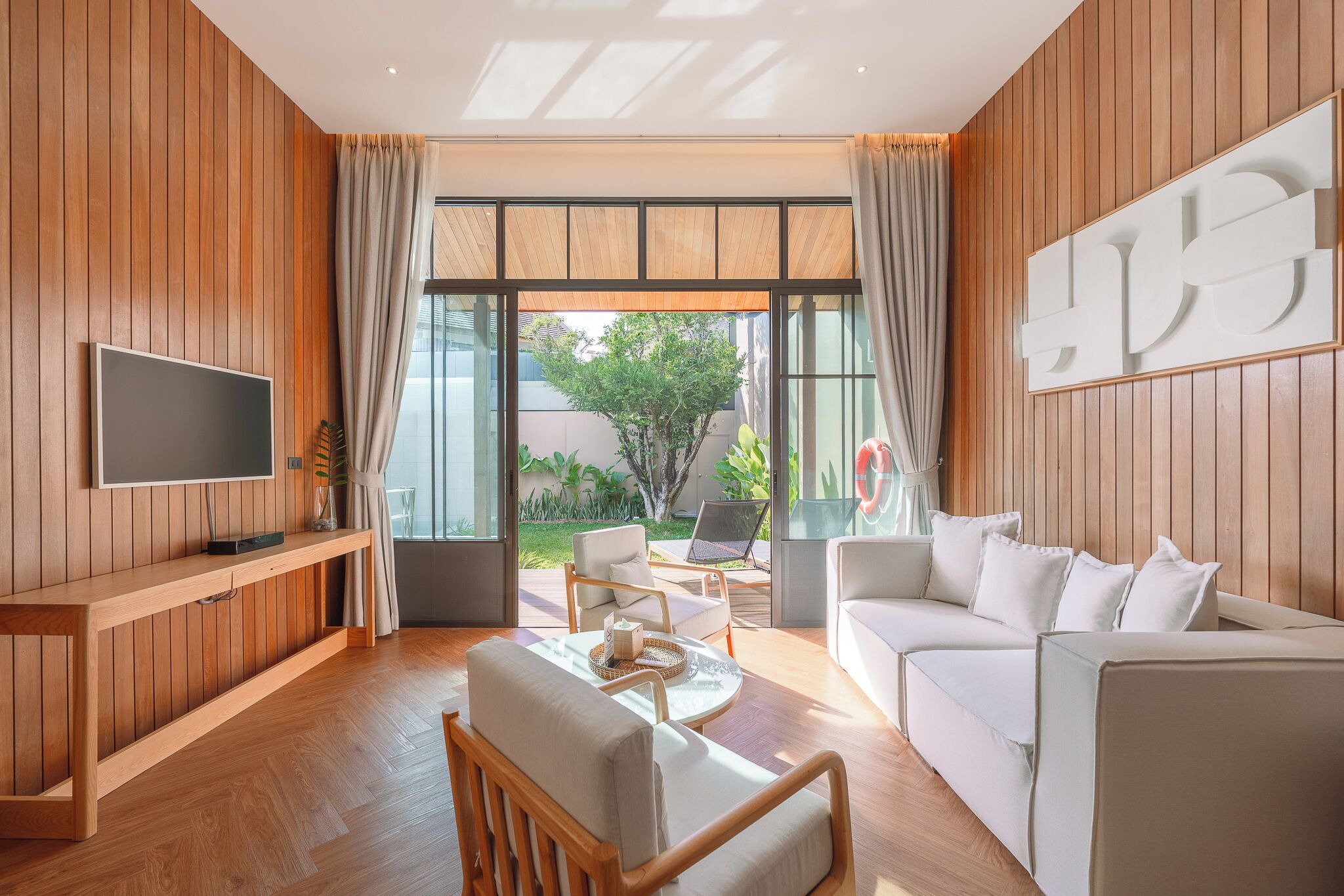 A modern living room with a flat-screen TV, wooden panel walls, a sofa, a coffee table, and a view of the outdoors.