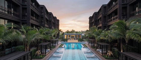 A modern hotel with a central swimming pool, surrounded by palm trees and lounge areas.