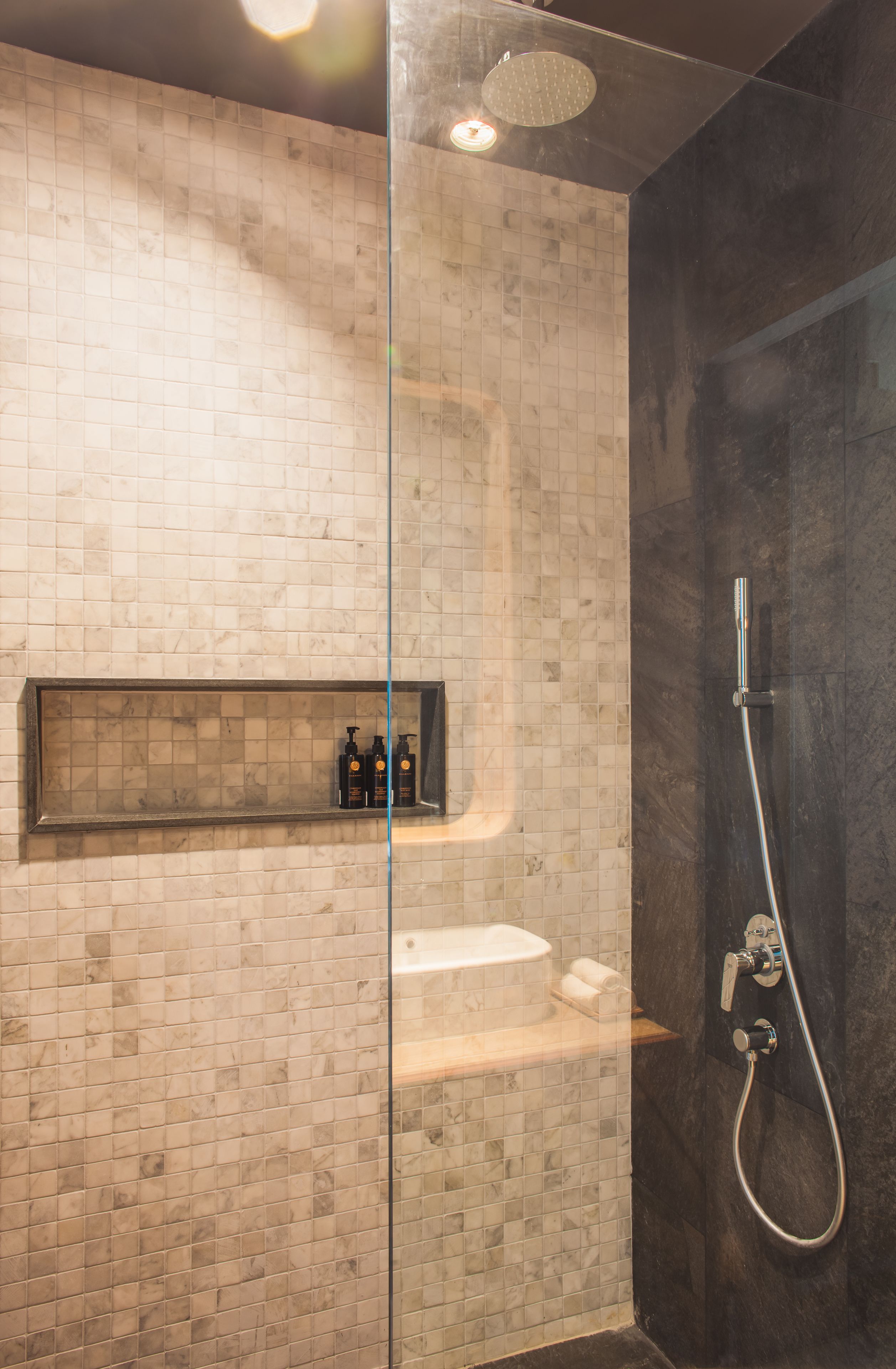 A modern bathroom with a glass partition, a shelf with toiletries, a shower area, and a marble-tiled wall.