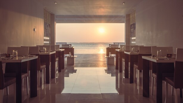 Un restaurant offrant une vue imprenable sur le coucher de soleil sur l’eau, avec des tables et des chaises au design moderne, et un sol épuré et réfléchissant.