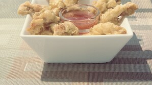 Un bol carré blanc rempli de morceaux de poulet frit et un petit récipient en verre contenant de la sauce, le tout posé sur une table recouverte d’un set de table en fibres tressées.