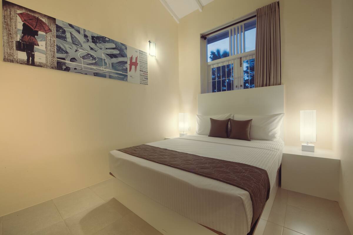 A neatly arranged bedroom with a large bed, a window with curtains, and a wall-mounted light fixture.