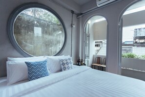 A bedroom with a large round window, a bed with white bedding and patterned pillows, a small table with bottles, and a view of the outside through an arched doorway. - A Local Something Hostel (Trang)
