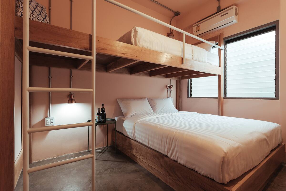 A bunk bed with a wooden frame and white bedding, a bedside table with bottles, and a window with blinds.