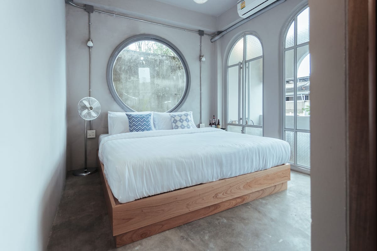 A modern bedroom with a large round window, a standing fan, and a wooden bed frame.