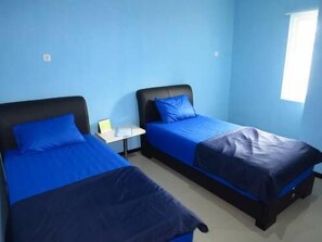 A room with two beds, each with a blue bedspread and pillow. There is a small table between the beds with a laptop and a lamp. The walls are painted blue, and there is a window on the right side. - Sleep Inn Pattaya - Hostel (Pattaya)