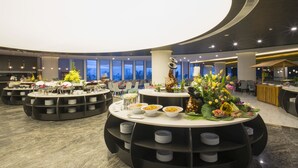 Un moderno vestíbulo de hotel con mesas redondas, estaciones de buffet y vistas a la ciudad a través de amplias ventanas.