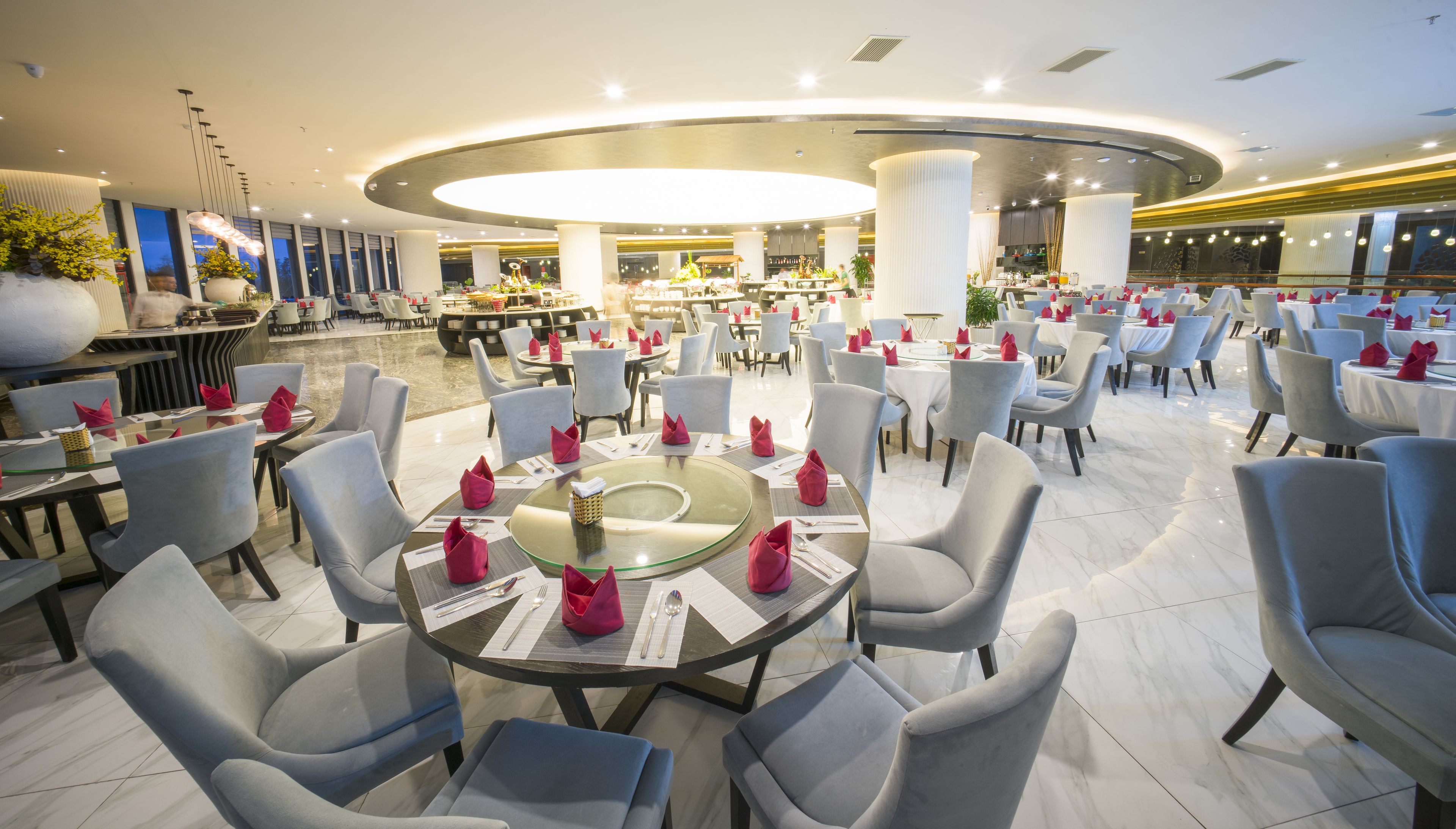 Un restaurant spacieux avec des tables rondes et des chaises grises, où l’on trouve une table centrale en verre, agrémentée de serviettes rouges et de couverts.