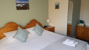 Una cama bien hecha con colcha blanca y dos almohadas, una mesita de noche con lámpara, un aparador con espejo y dos cuadros en la pared.