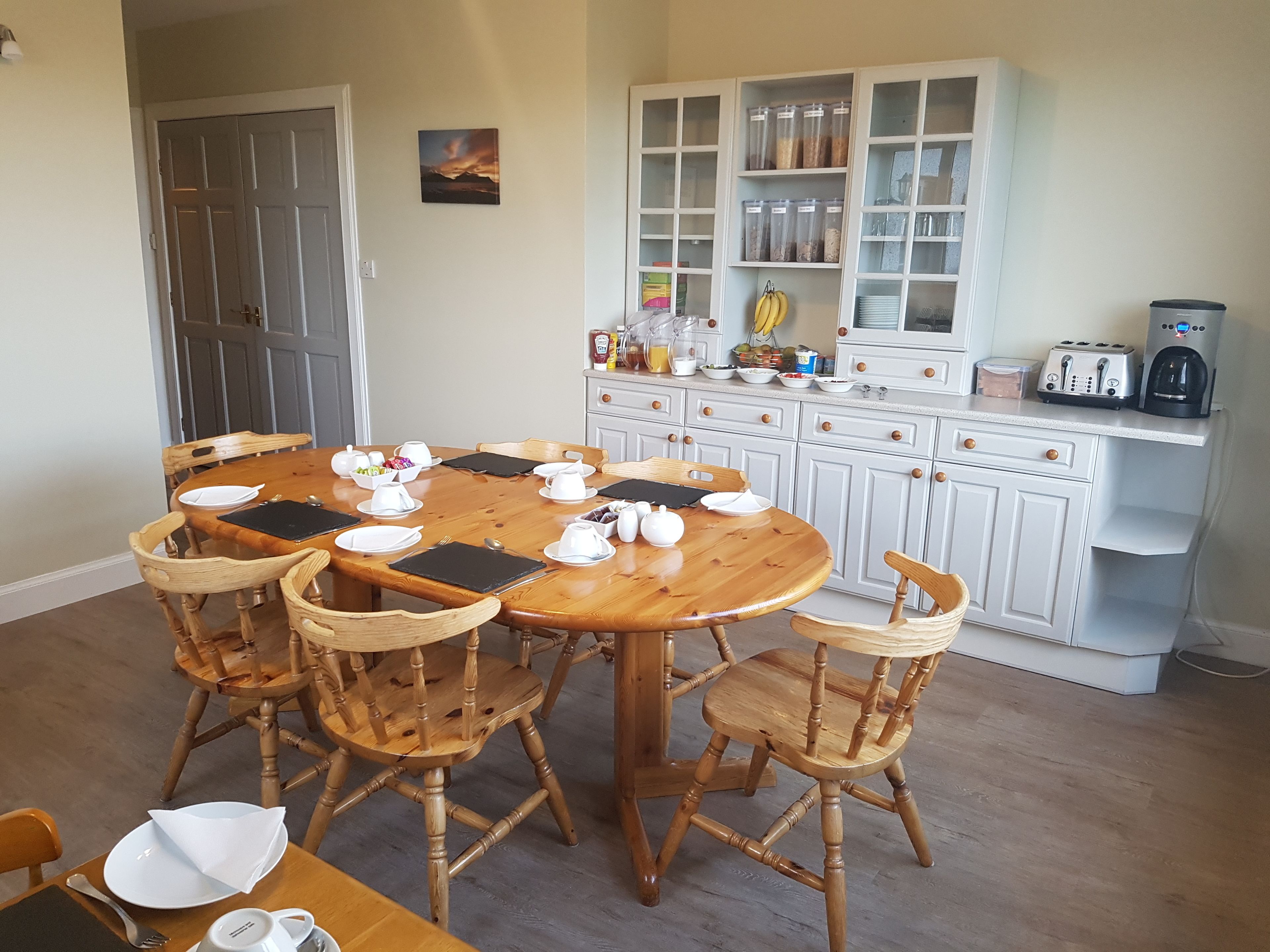 A dining area with a wooden table and chairs, a kitchen with white cabinets and a countertop, and a door in the background.