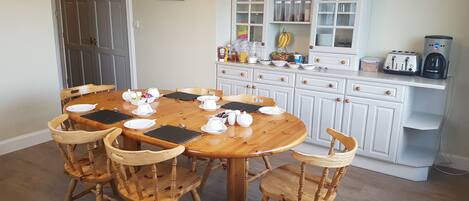 Un'area pranzo con tavolo e sedie in legno, una cucina con mobili bianchi e un piano di lavoro, e una porta sullo sfondo.