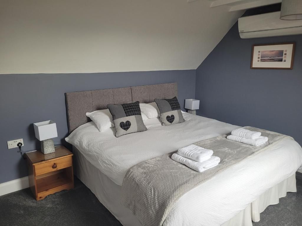 Una cama cuidadosamente tendida con tablero de cabecera gris, ropa de cama blanca y almohadas con estampado de corazones. Junto a la cama, una mesita de noche de madera con una lámpara y un pequeño adorno.