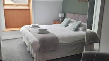 Un dormitorio con cama, una silla, una mesita de noche, una lámpara y una ventana.