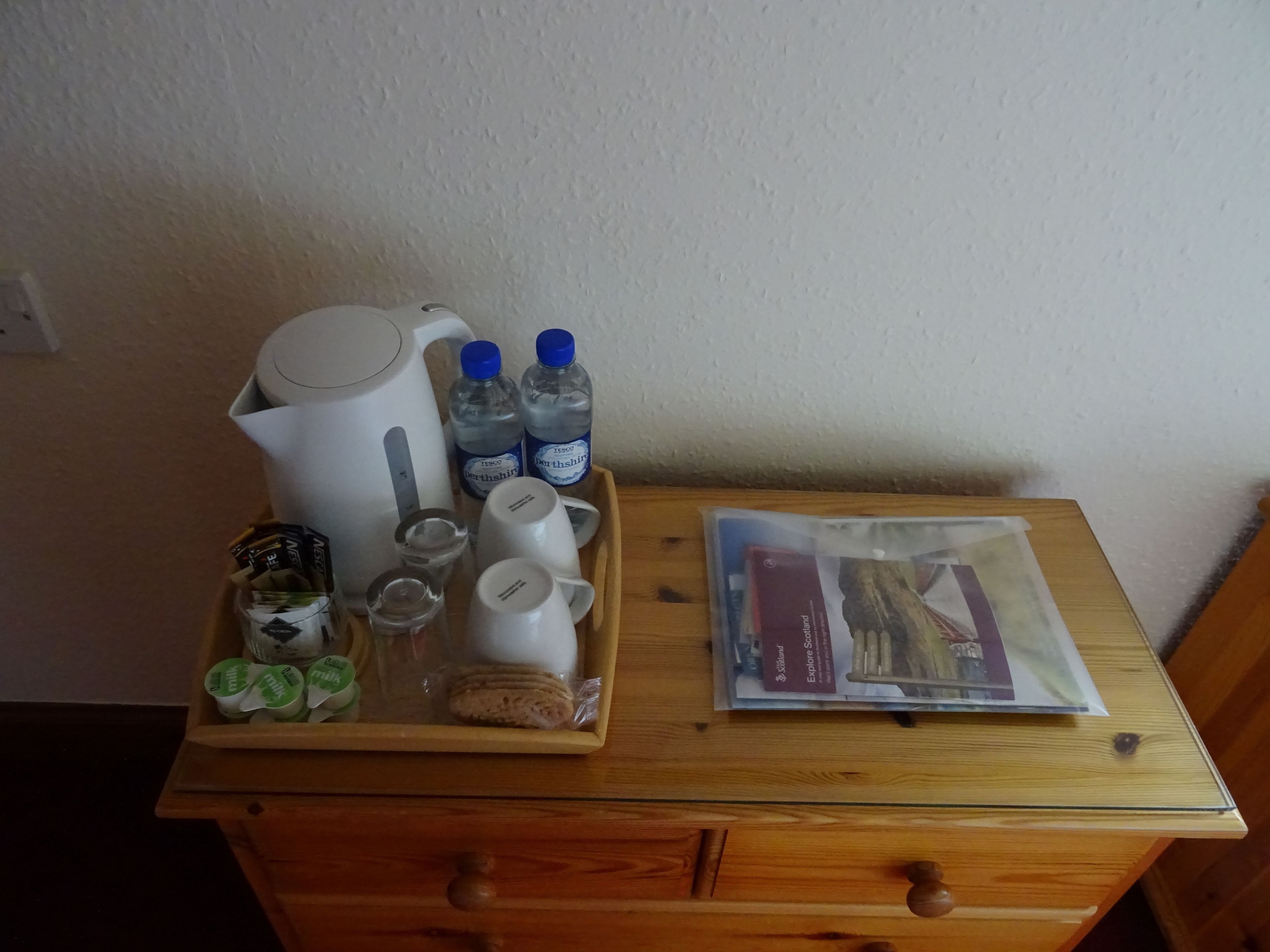 Une table en bois sur laquelle se trouve un plateau contenant une bouilloire, deux bouteilles d’eau, une tasse, un verre, un pot de yaourt et un paquet de biscuits.