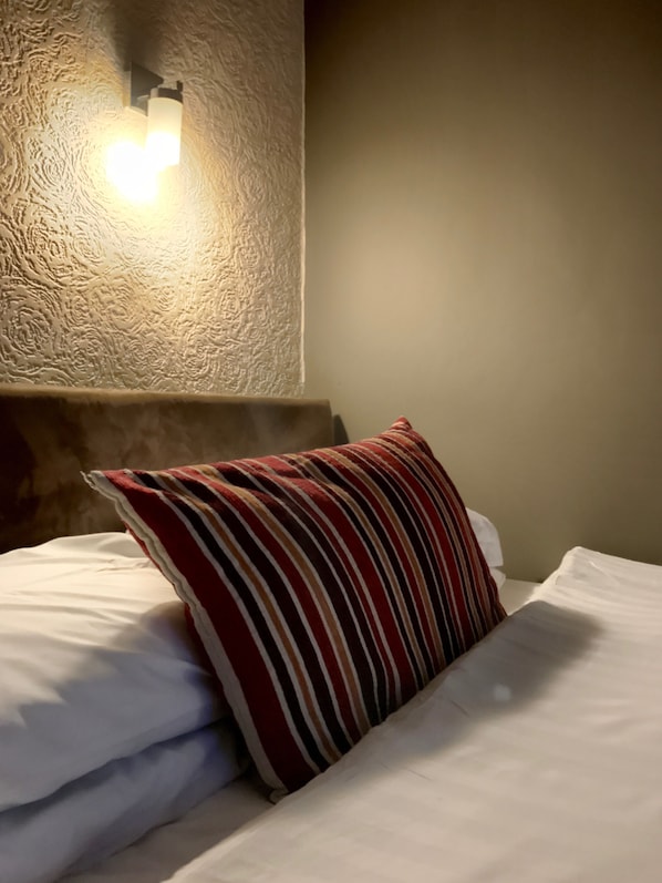 A neatly made bed with white linens and a striped pillow, a textured wall, and a wall-mounted light fixture. - Glenlossie Guest House (Dumfries)