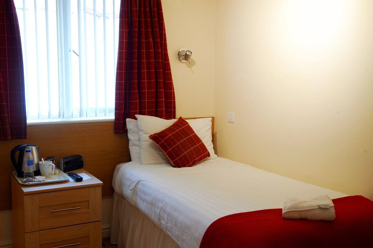Ein Hotelzimmer mit einem Bett, einem Nachttisch, einem Fenster mit Vorhängen, einer Wandleuchte und einer roten Decke auf dem Bett.