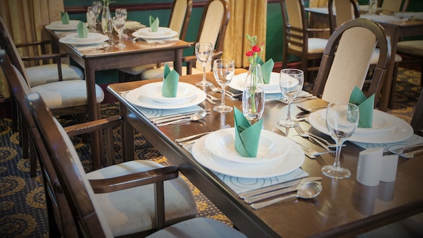 Une salle à manger élégante, avec plusieurs tables dressées pour le service, chacune agrémentée d’assiettes blanches, de serviettes vertes et de verres transparents.