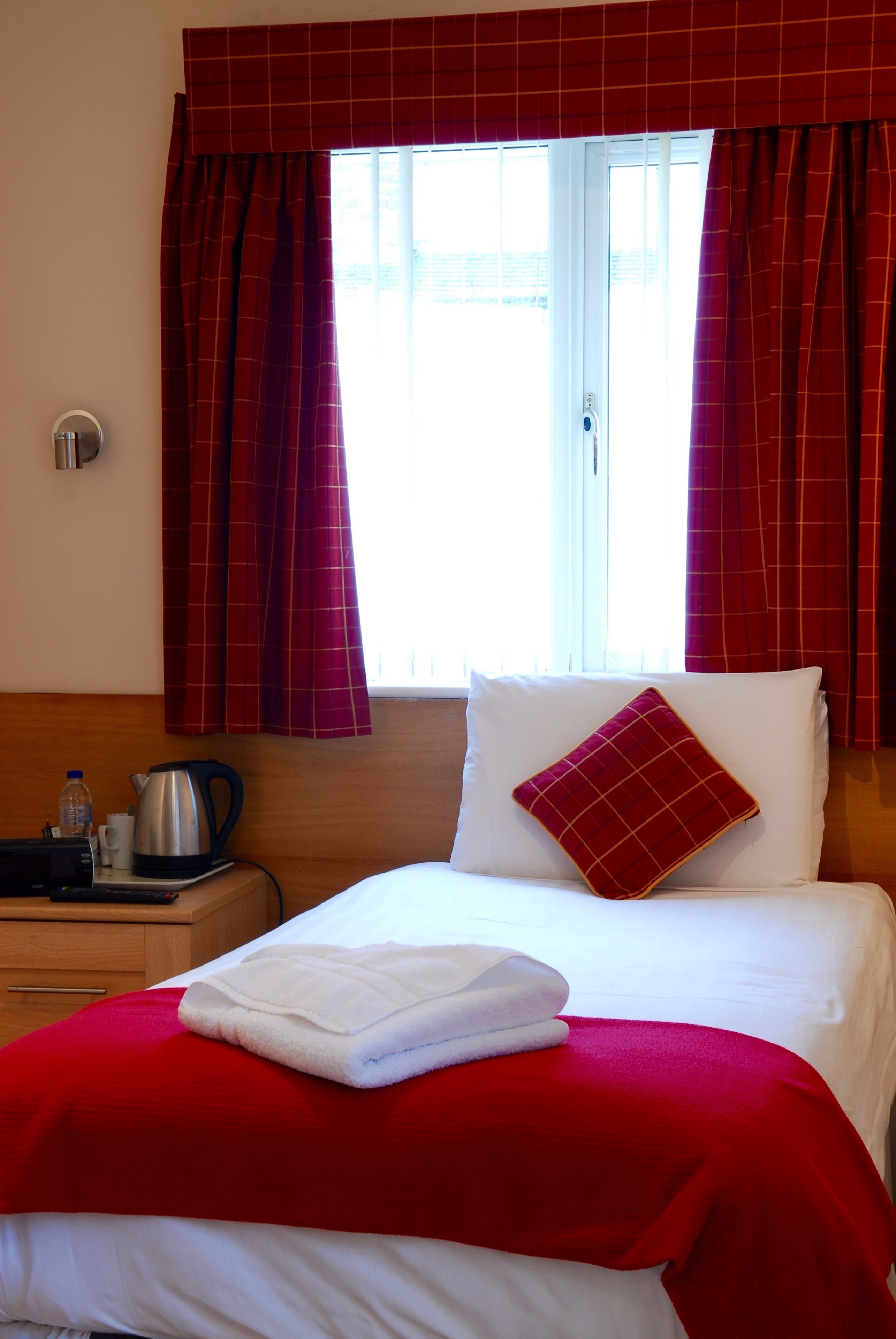 A hotel room with a bed, red curtains, a window, a bedside table with a kettle, and a red throw blanket.