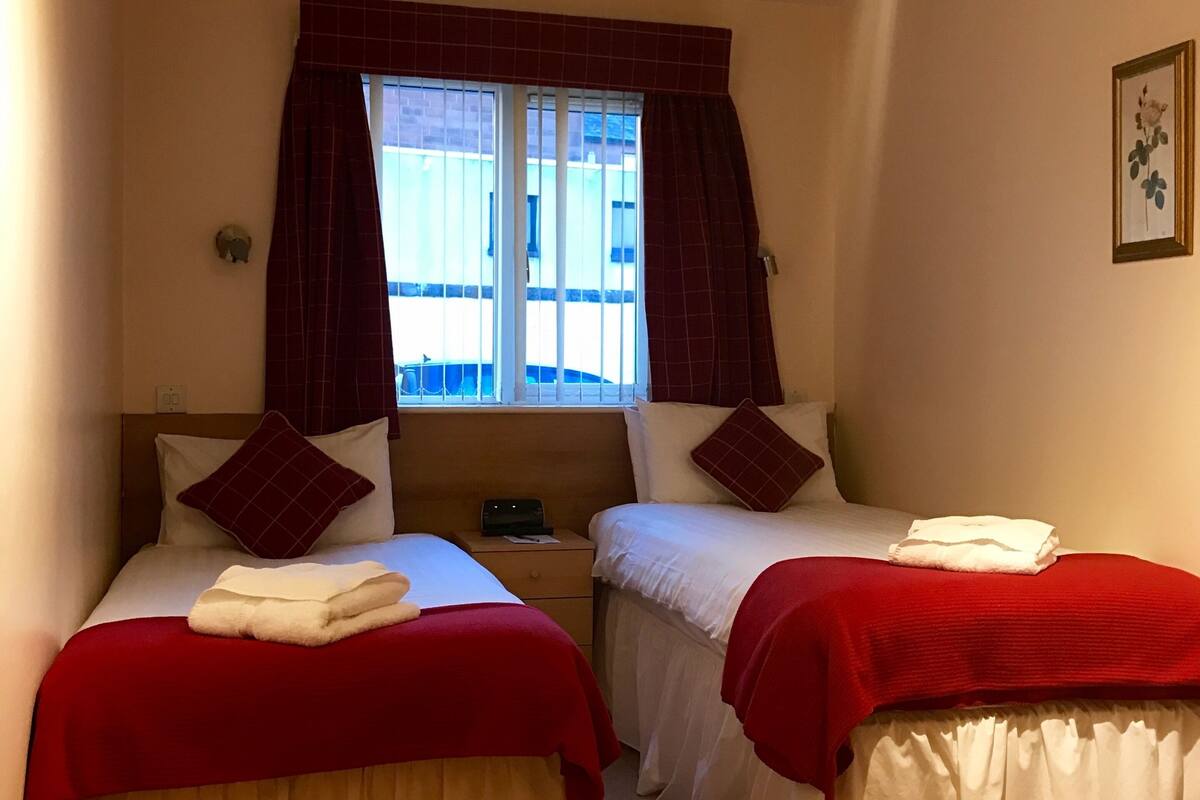 Ein Hotelzimmer mit zwei Betten, jedes mit roter Decke und Kissen, einem Fenster mit Vorhängen, einem kleinen Nachttisch und einem gerahmten Bild an der Wand.