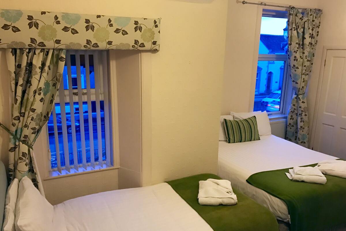 Ein Hotelzimmer mit zwei Betten, jeweils mit einer grünen Tagesdecke und weißen Bettwäsche. Ein Fenster bietet einen Blick auf ein Gebäude.