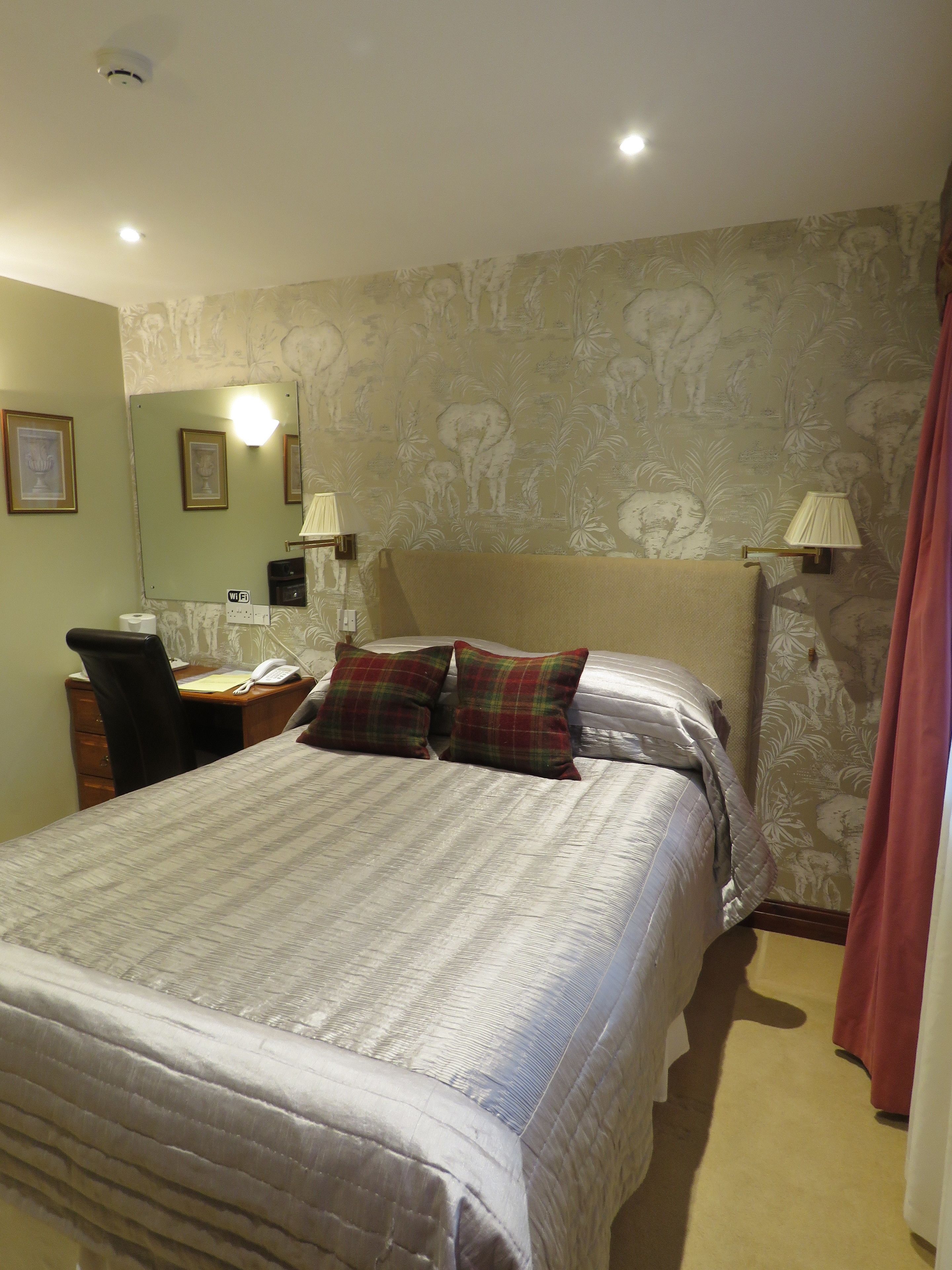 A neatly made bed with a patterned bedspread, a desk with a telephone and papers, a chair, and a wall with a floral wallpaper pattern.