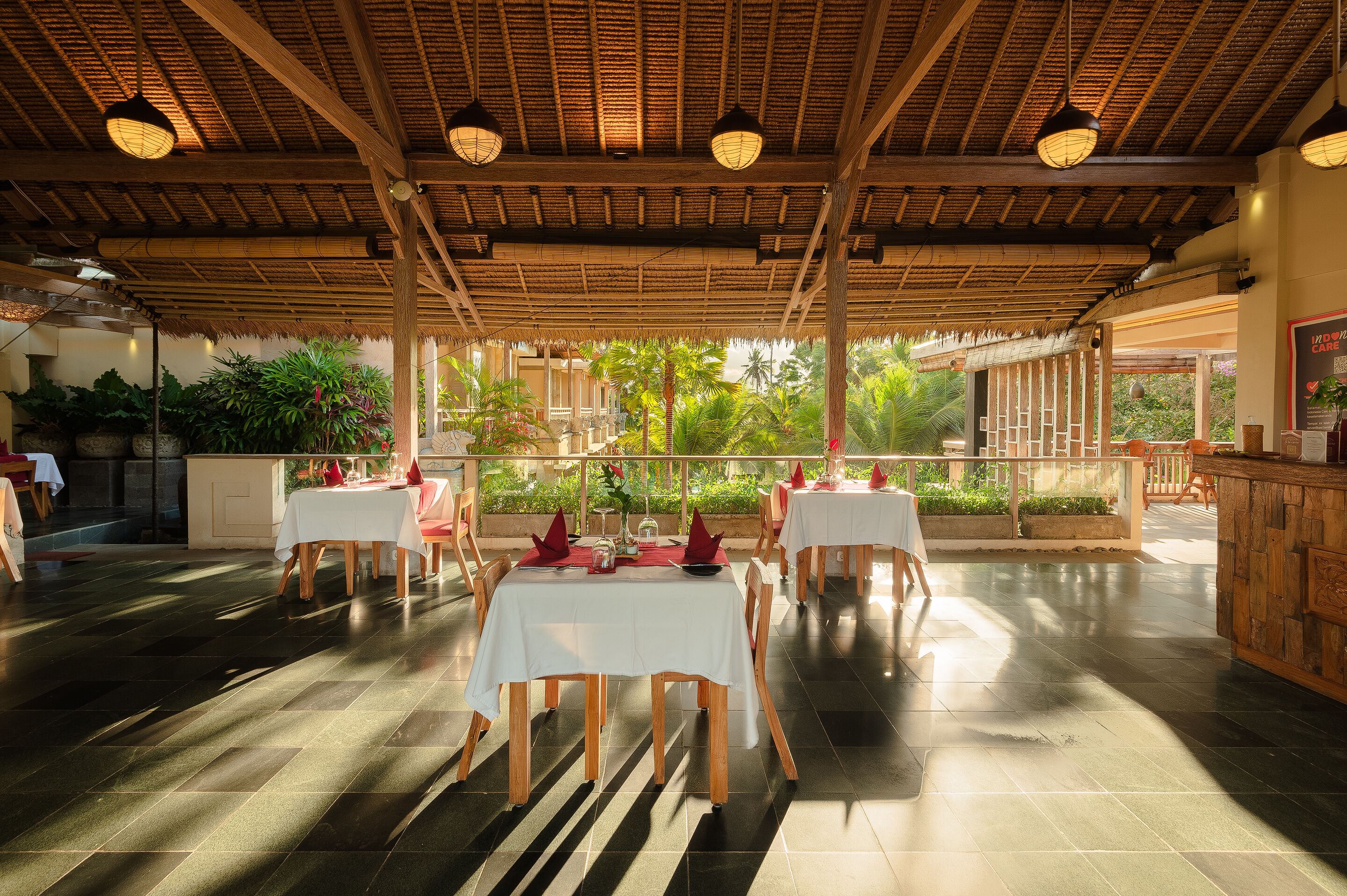 Un área de comedor al aire libre con mesas preparadas para dos, cubierta por un techo de madera con luces colgantes, rodeada de abundante follaje.