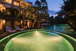 A pool area with lounge chairs and umbrellas, surrounded by lush greenery and a building with multiple floors. - The Udaya Resorts and Spa (Ubud)