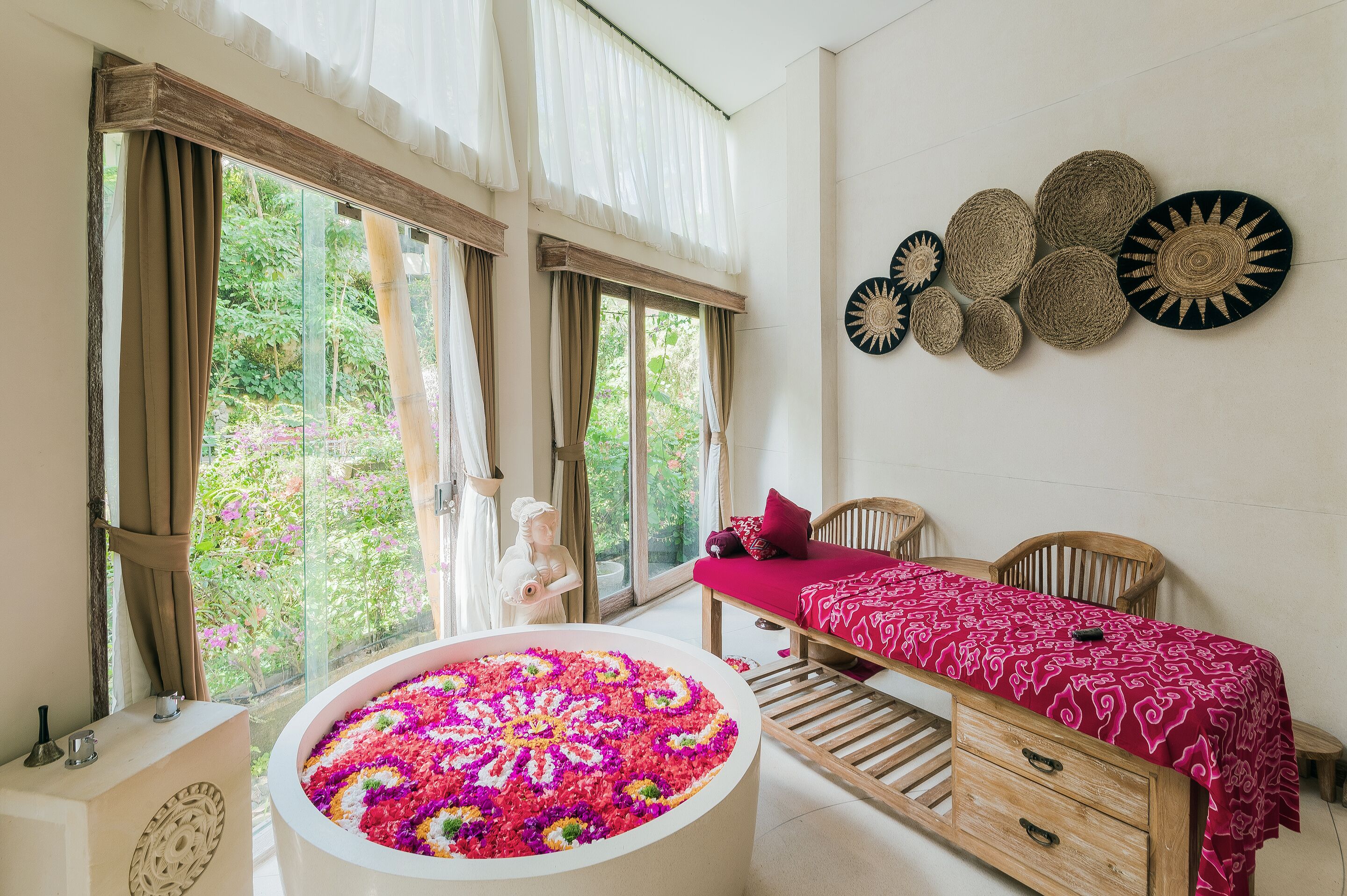 A room with a large window, a bathtub with flower petals, a bed with a red bedspread, and wall decor.