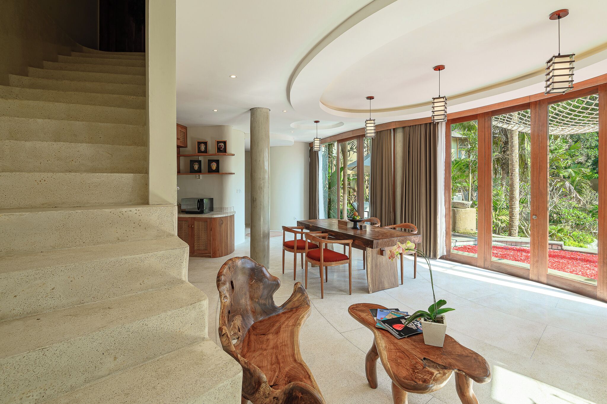 A spacious living area with a wooden staircase, a dining table with chairs, and large windows offering a view of greenery.