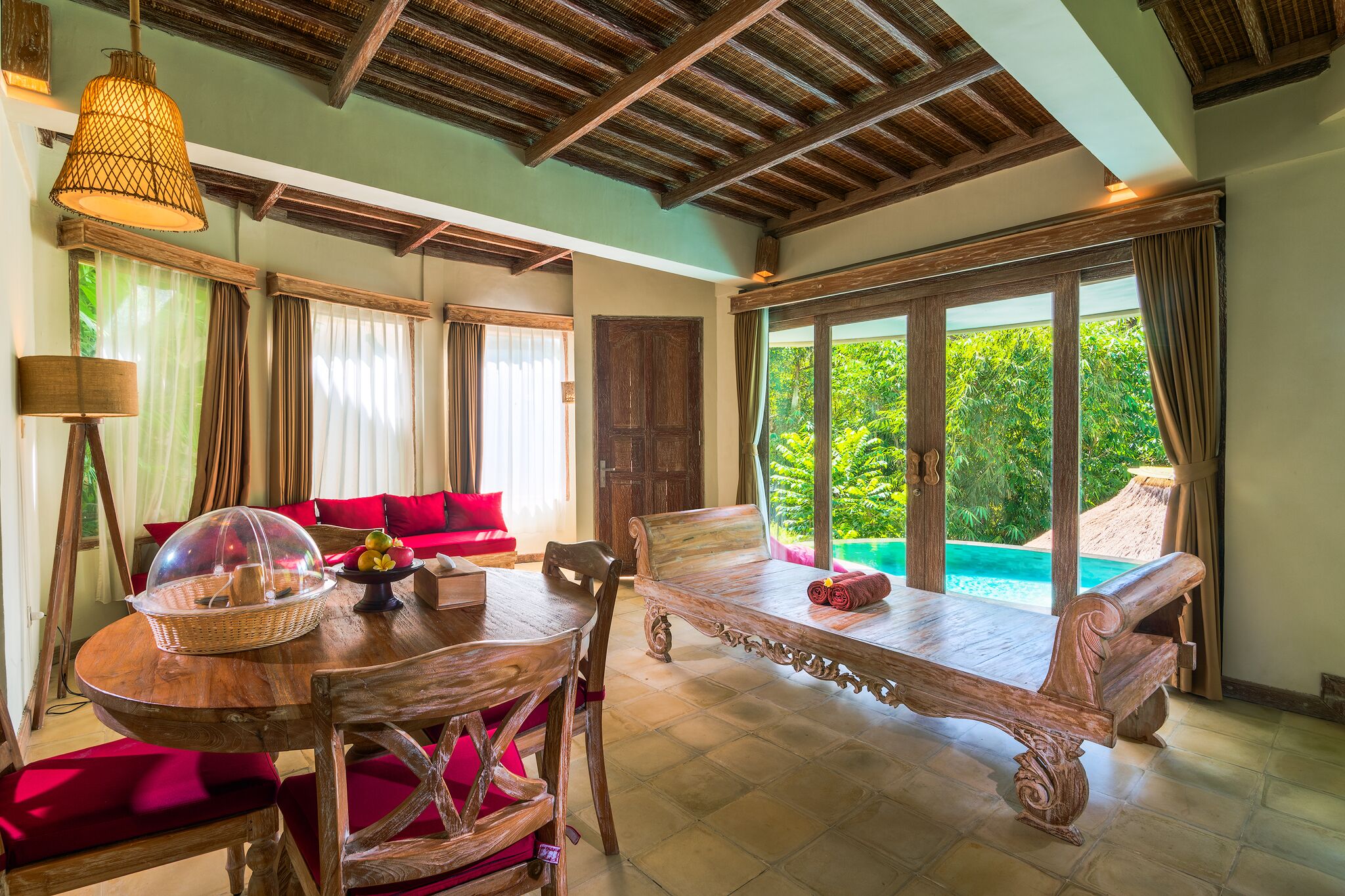 A spacious room with a wooden dining table, red upholstered chairs, a wooden bench, and a large window overlooking a pool.