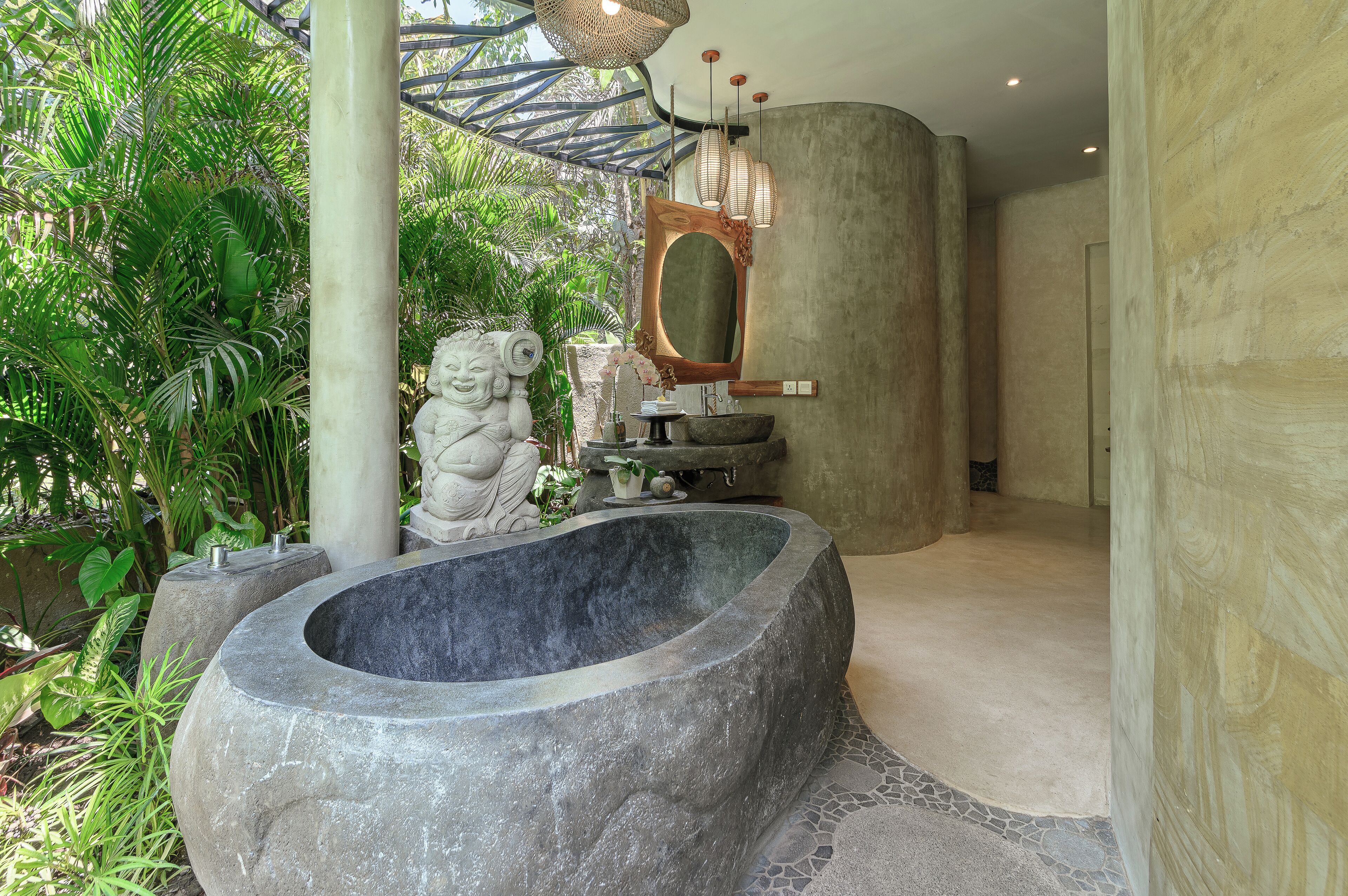 A stone bathtub in a lush, green bathroom with a large mirror and a statue.