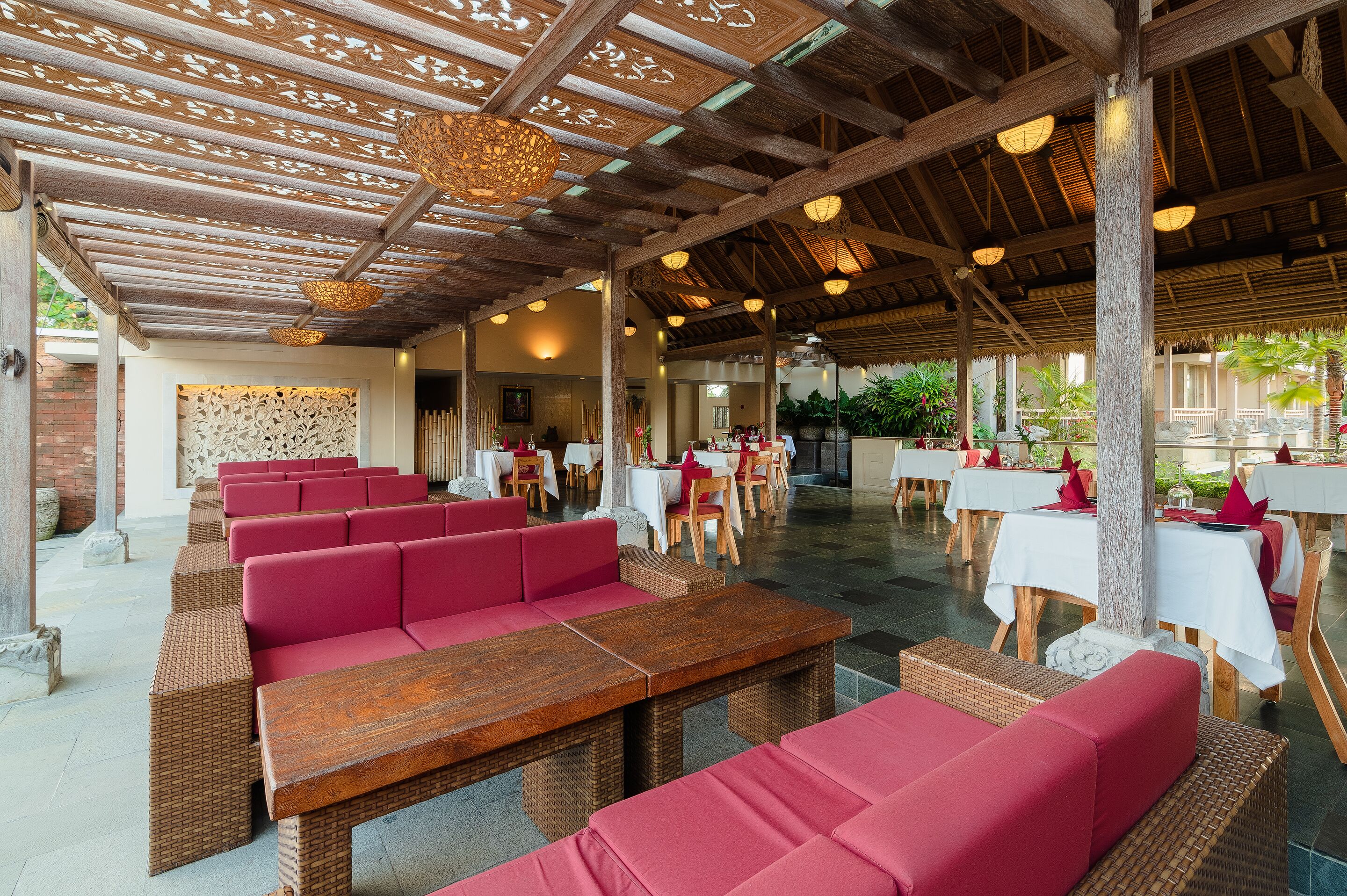 Un espace extérieur aménagé, abrité et doté de mobilier en osier et de coussins rouges, de tables en bois et d’un plafond décoratif.