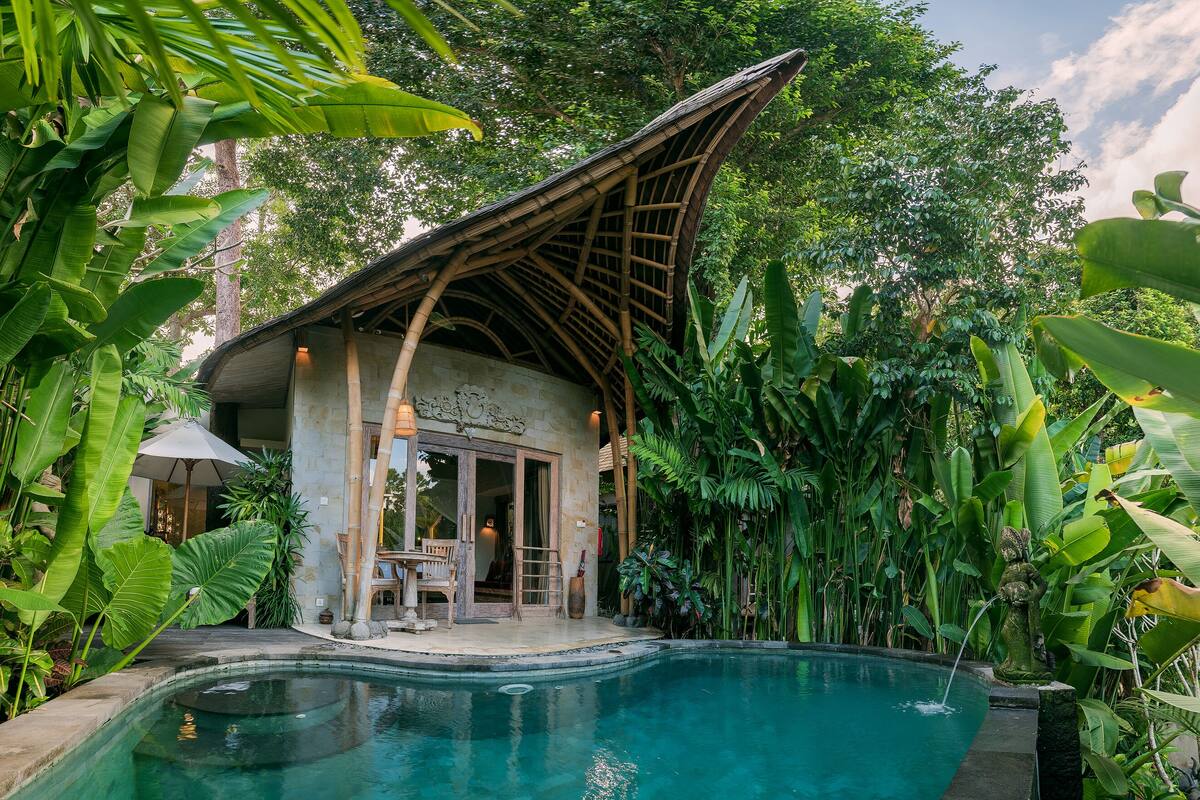 Un área junto a la piscina con una estructura de bambú, un edificio de piedra y abundante vegetación.