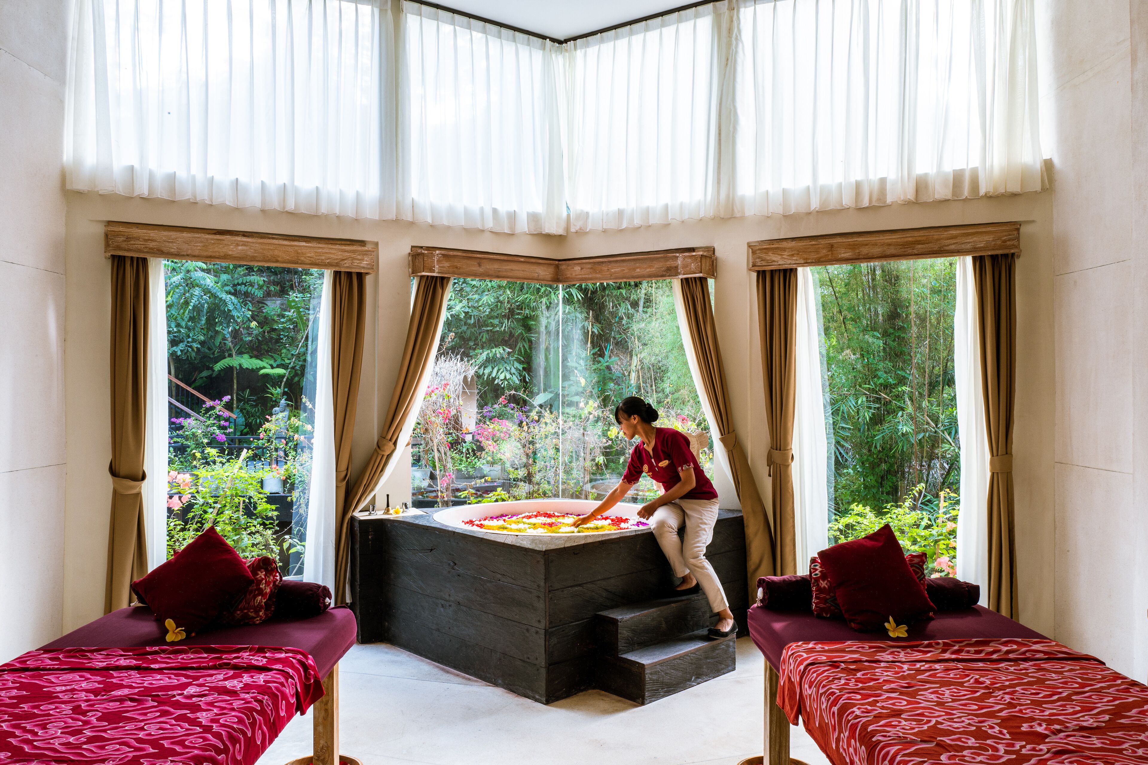 A room with a large bathtub, two beds with red bedding, and large windows with curtains.