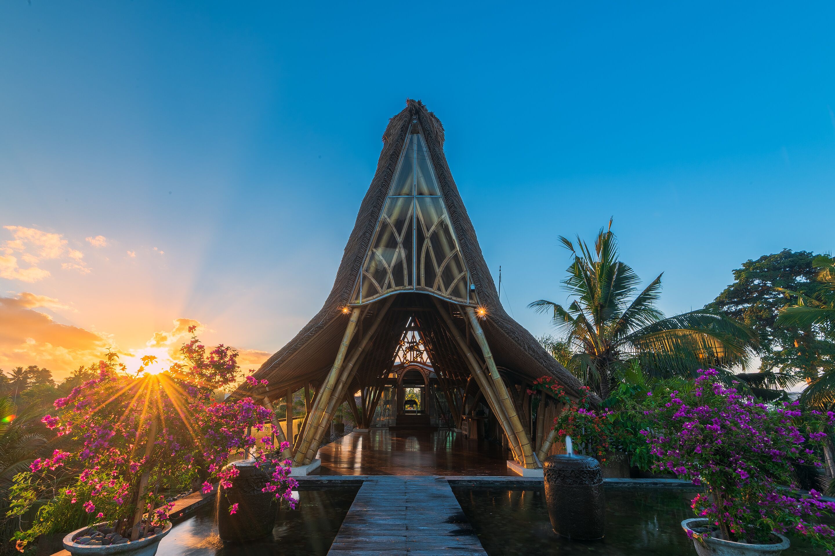 Un grand pavillon ouvert, doté d’une arche centrale, entouré d’une végétation tropicale luxuriante et agrémenté d’un bassin d’eau dont la surface réfléchit la lumière.