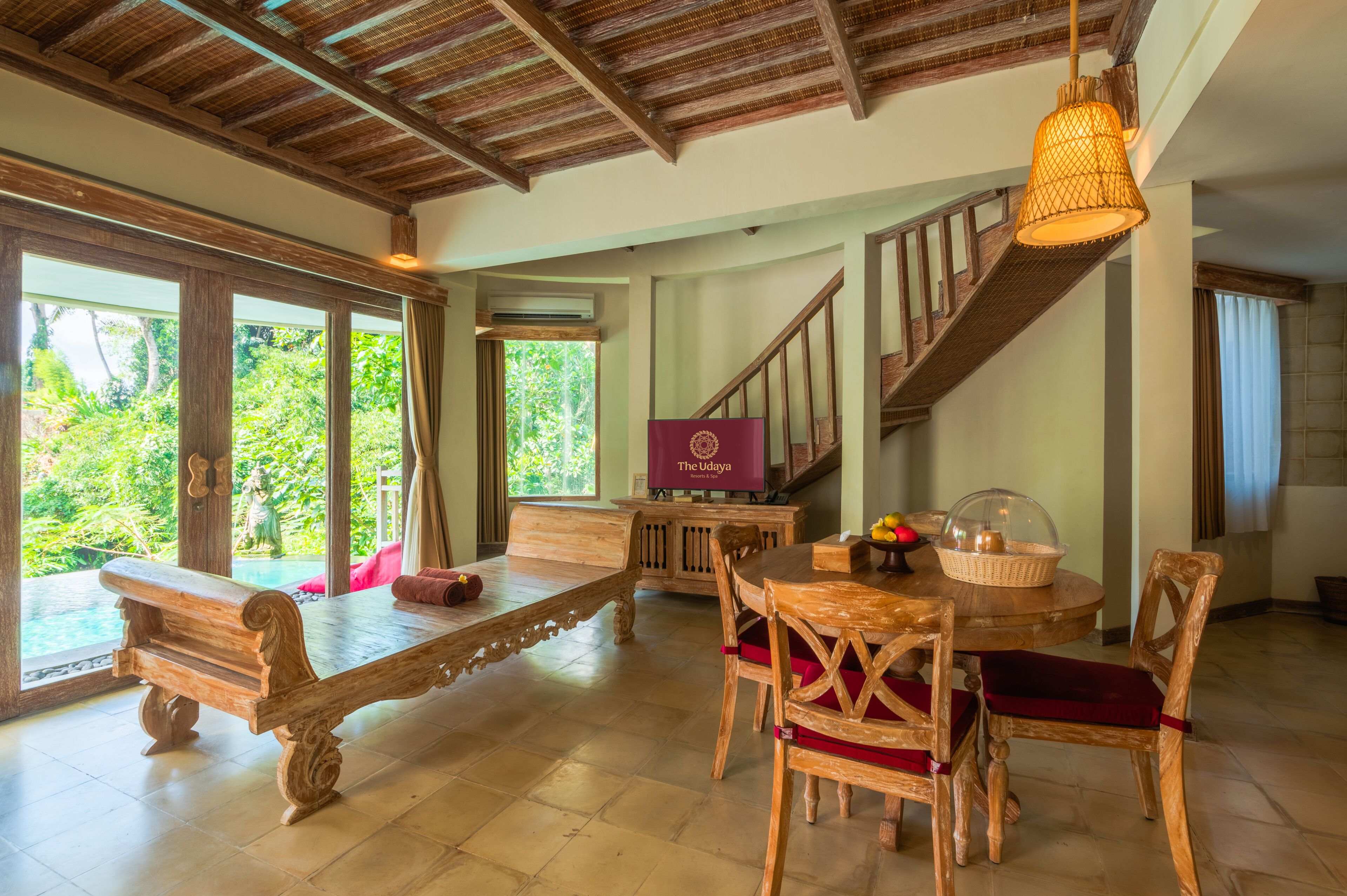 A spacious room with a wooden bench, a dining table with chairs, a staircase, and a large window offering a view of greenery.