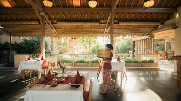 An indoor dining area with tables set for a meal, a person carrying a basket, and a view of greenery outside.