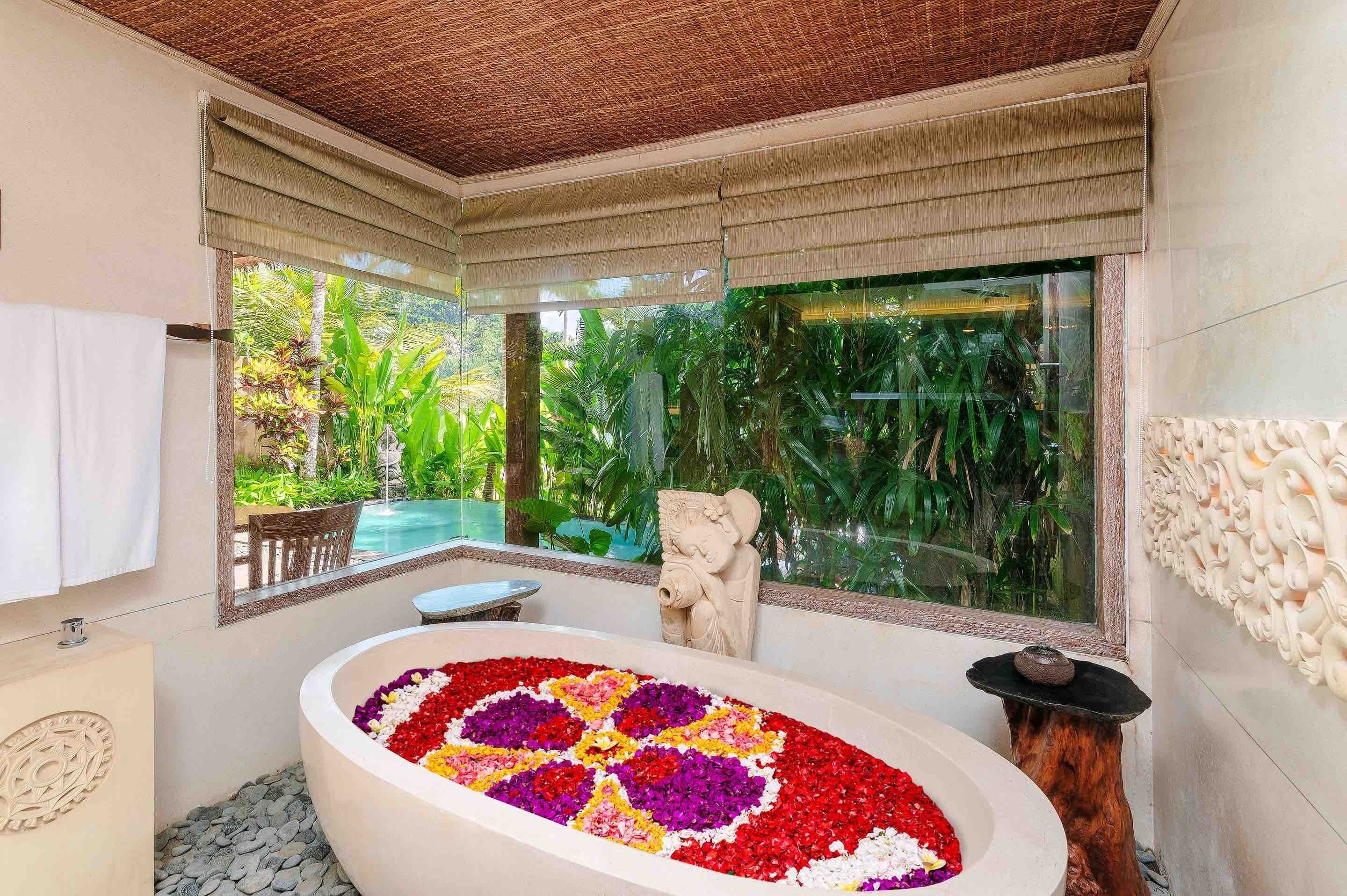 A bathroom with a large bathtub surrounded by a flower arrangement, a stone sculpture, and a view of lush greenery.