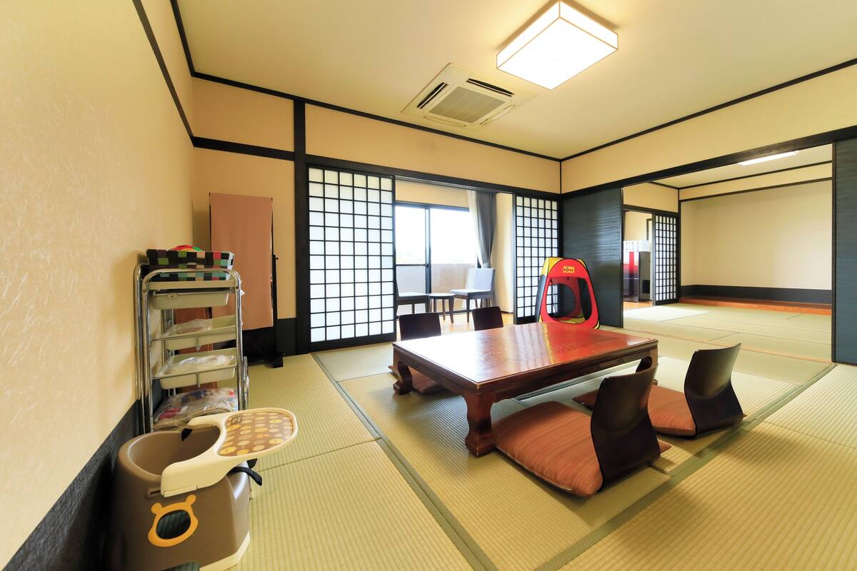 A tatami mat room with a wooden table, chairs, a red chair, a trash bin, and a shelf with items.