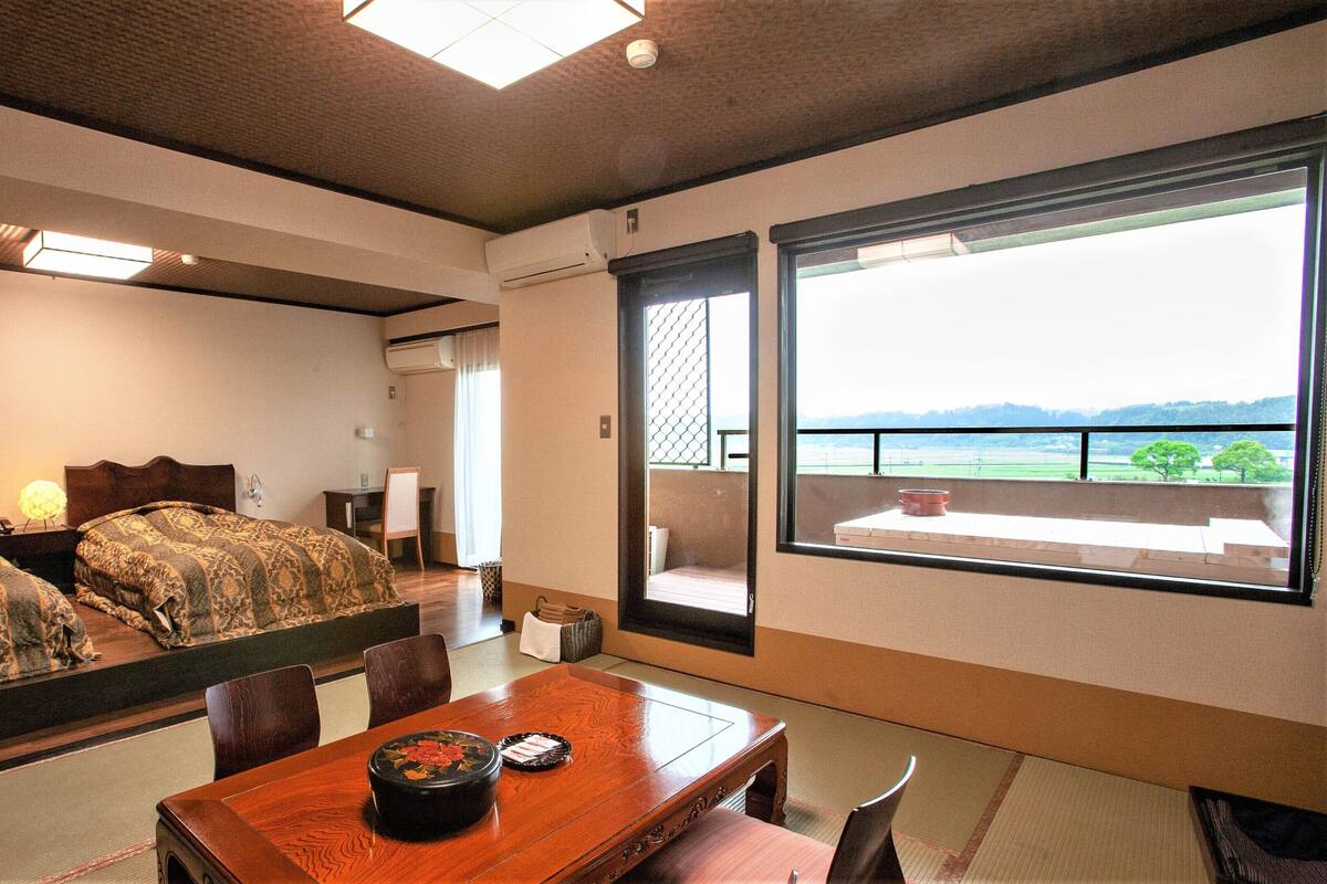 A hotel room with a bed, a dining table with a traditional Japanese tea set, a view of a balcony with a railing, and an air conditioning unit on the wall.