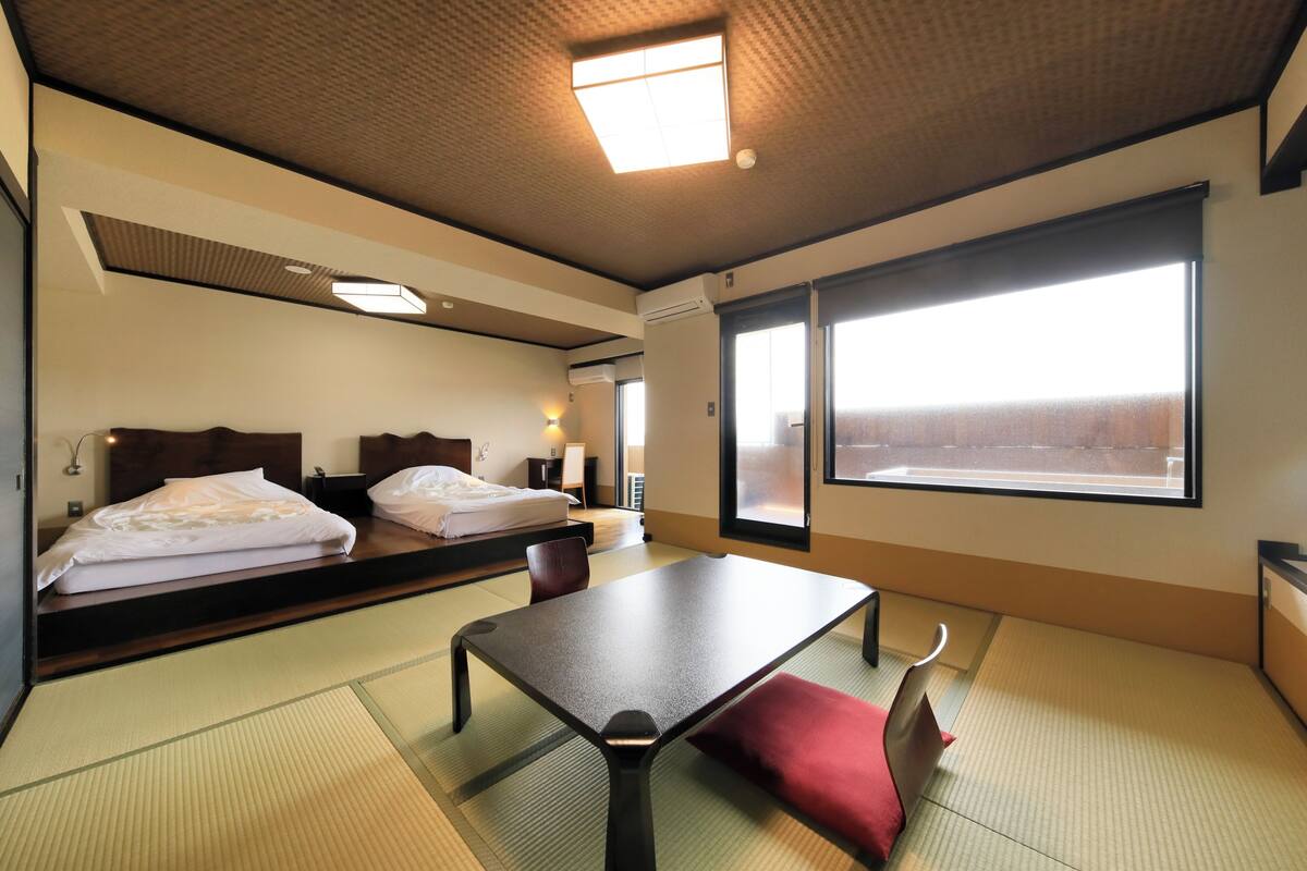 A hotel room with tatami flooring, a bed with a white comforter, a table with a red cushion, and a large window with a view.