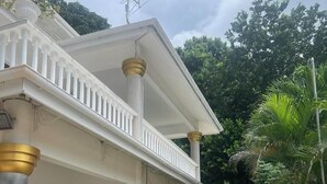 Una casa de dos pisos con balcón, rodeada de exuberante vegetación y un césped bien cuidado.