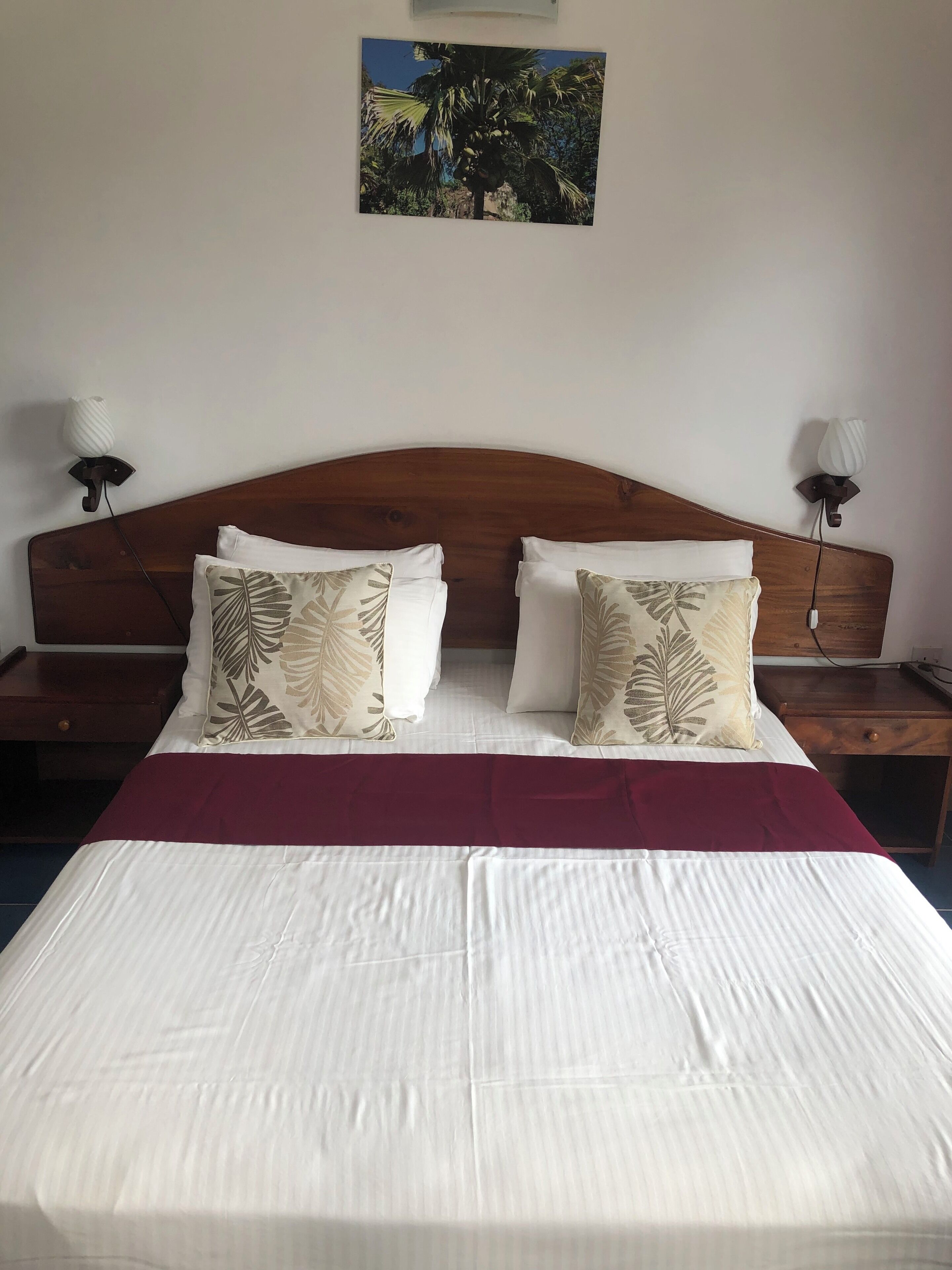 A neatly made bed with a wooden headboard, two bedside tables with lamps, and a tropical-themed wall hanging.
